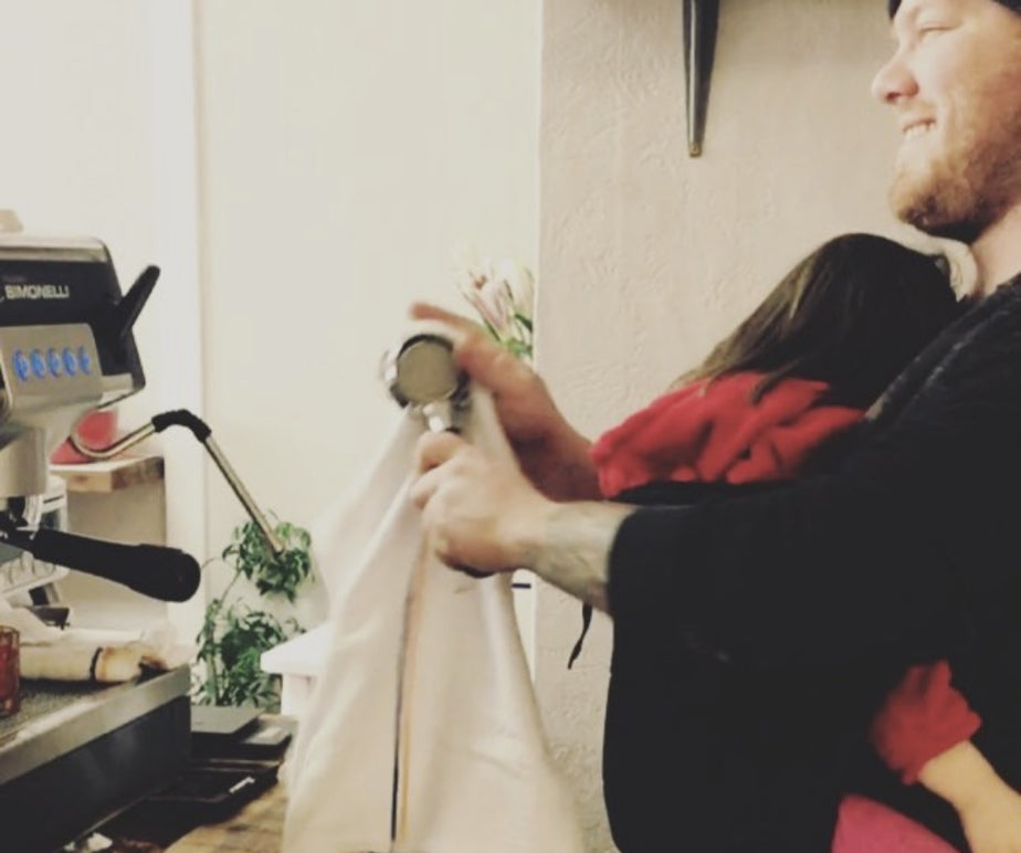 Braden making coffee holding his daughter in a carrier in a family moment.